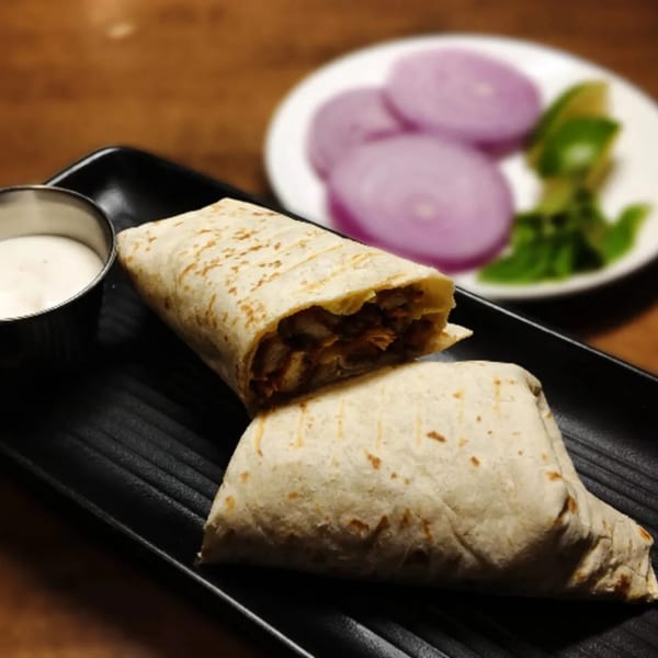 Two chicken shawarma wraps on a black plate with a side of sauce, red onions, and lime.