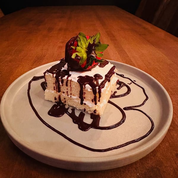 Slice of cake with white frosting, chocolate drizzle, and a chocolate-covered strawberry on a plate.