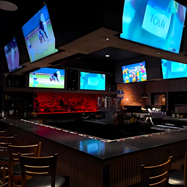 A dark, modern bar with multiple large screens showing sports and entertainment, and a red-lit back wall.